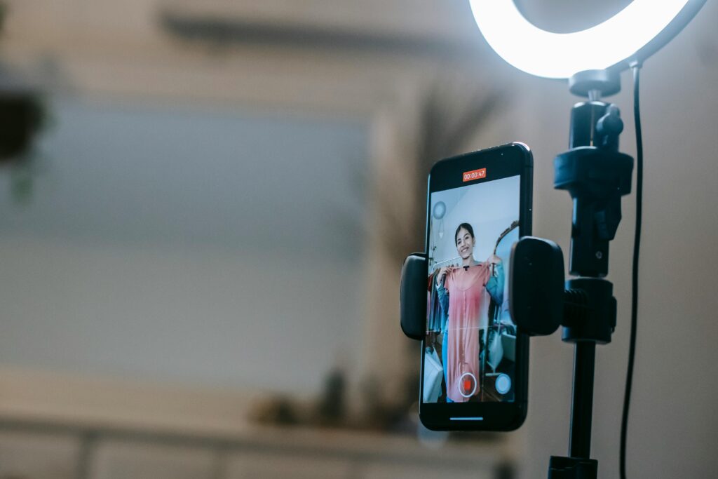 Anonymous female blogger standing in room and shooting vlog on smartphone with ring lamp while demonstrating pink dress in hands