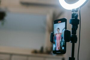 Anonymous female blogger standing in room and shooting vlog on smartphone with ring lamp while demonstrating pink dress in hands